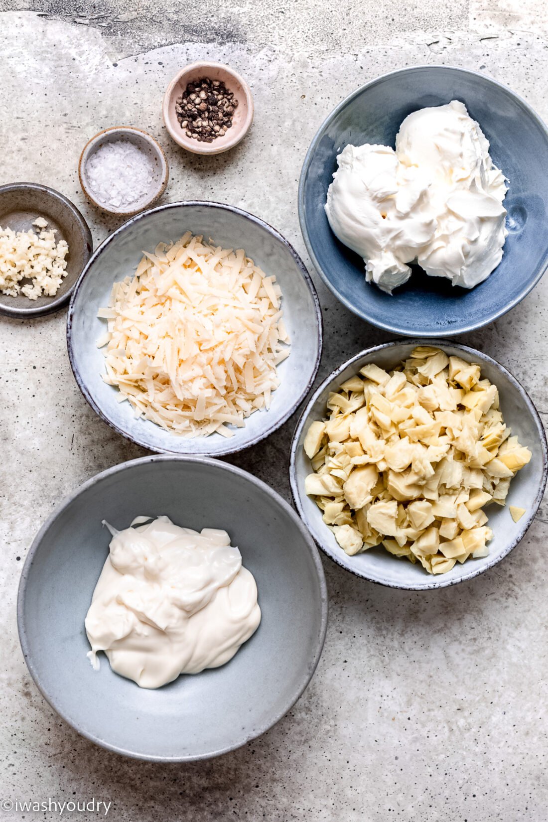 Hot artichoke dip ingredients in bowls on marble countertop. 