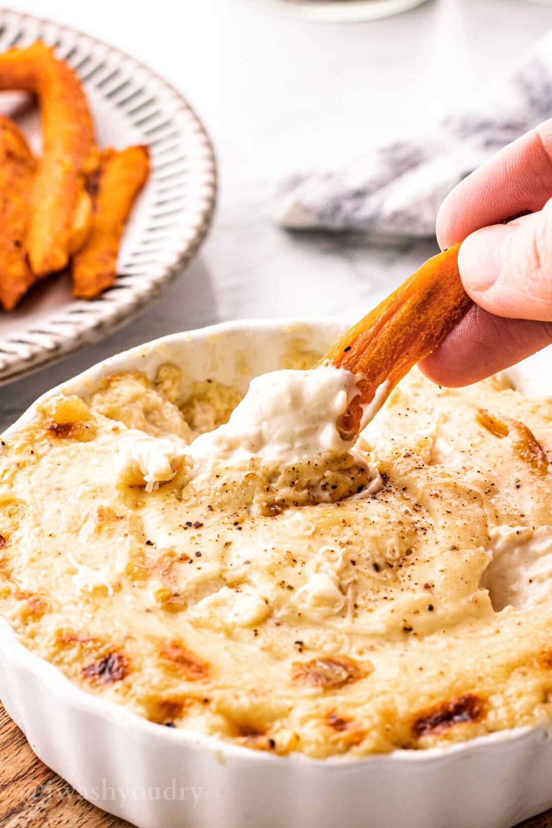 Sweet potato wedge dipping into roasted garlic dip in white dish.