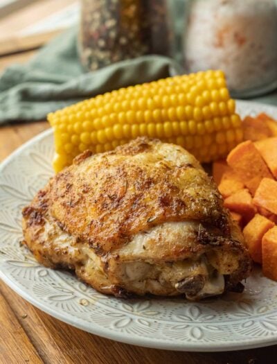Cooked chicken thigh on white plate with corn and sweet potato.